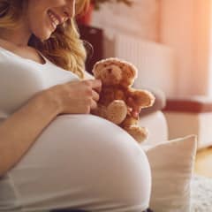 Pregnant Woman Holding Teddy Bear and smiling, sitting on the floor in her living room. Wearing white T-shirt . Woman has brown curly hair and beautiful smile. Sunlight in background.