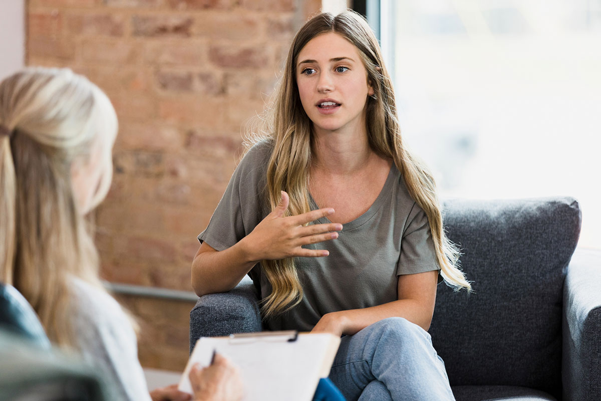 The young adult woman gestures as she talks to the unrecognizable female mature adult counselor.