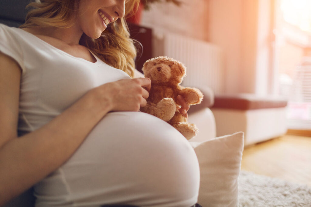 Pregnant Woman Holding Teddy Bear and smiling, sitting on the floor in her living room. Wearing white T-shirt . Woman has brown curly hair and beautiful smile. Sunlight in background.